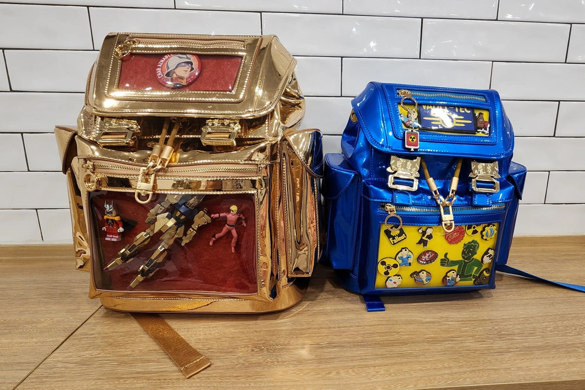 Two shiny Cyberpunk Ita Backpacks by Ita Bag Co sitting on a counter. On the left is a standard-size gold bag displaying a 1/144 Hyakushiki action figure on a red insert. On the right is a mini-size blue bag displaying an organized collection of gaming pins on a yellow insert.